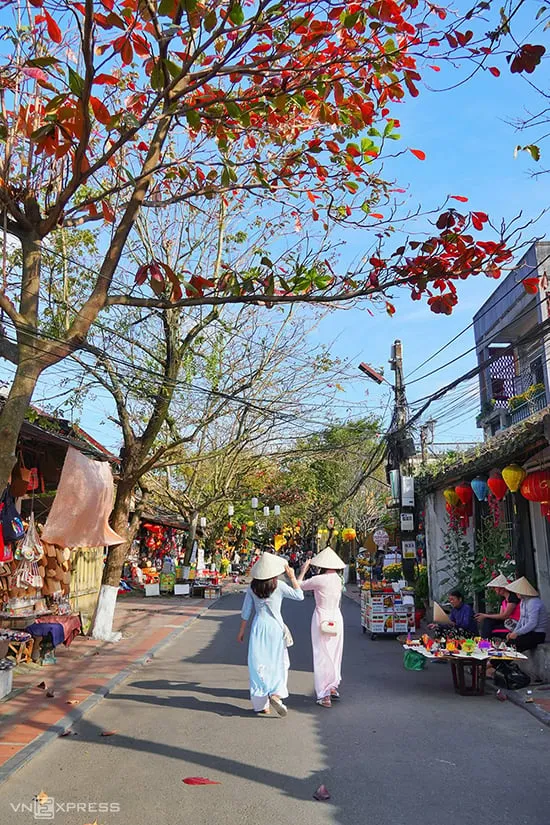 alt Hai người phụ nữ mặc áo dài đi trên đường Nguyễn Thị Minh Khai, Hội An, với hàng cây bàng lá đỏ hai bên đường.