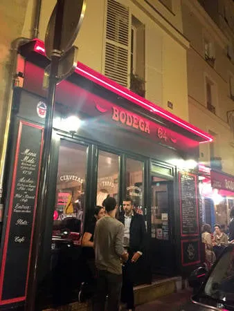 Alt: Khách hàng đứng uống bia và trò chuyện bên ngoài quán Bodega 64 ở Paris vào buổi tối