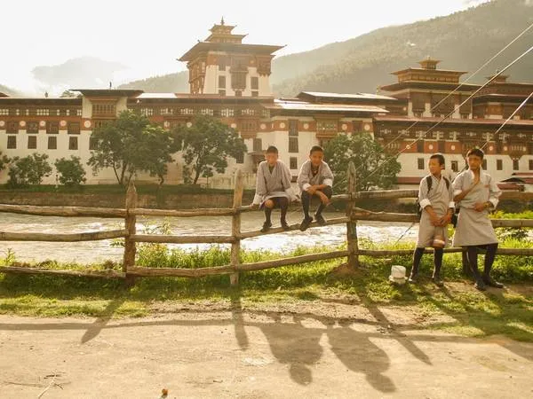 Học sinh Bhutan trong trang phục truyền thống đang đứng chờ xe buýt trước một Dzong, pháo đài kiêm tu viện, kiến trúc đặc trưng của Bhutan.