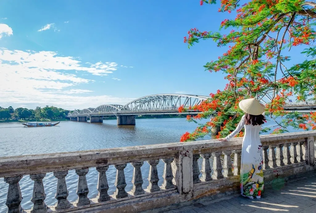 Phố đi bộ Nguyễn Đình Chiểu bên bờ sông Hương với hàng phượng vĩ đỏ rực, cầu Tràng Tiền ở phía xa và dòng người tản bộ nhộn nhịp.